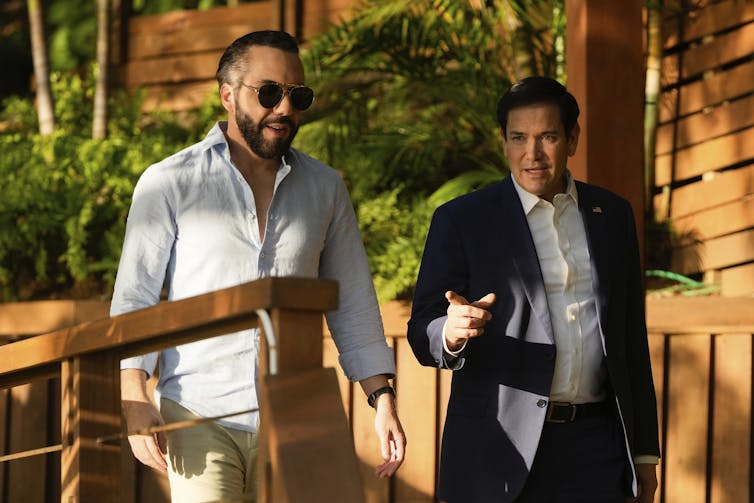 Two men walking and talking, one in a white shirt with sunglasses, the other in a suit.