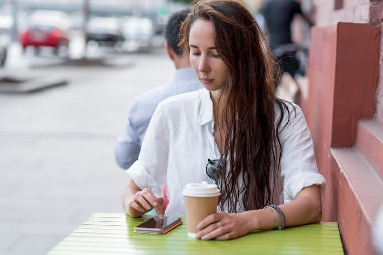 A young woman uses a cellphone while having coffee on an outdoor patio